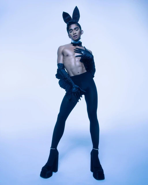 An adult male poses in a studio wearing a bunny costume with high platform boots, black tights, and a bow tie against a light blue background.