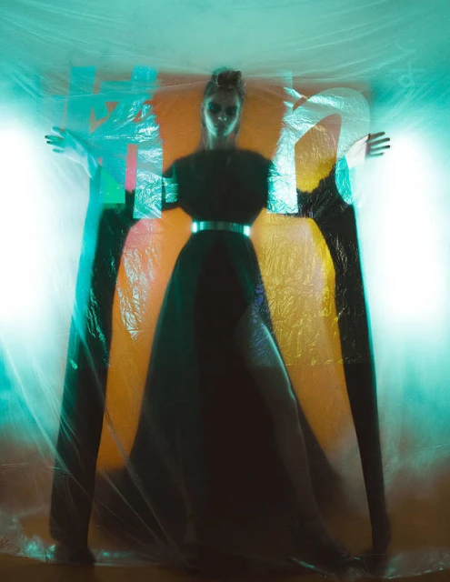 Woman in black dress posing with arms outstretched inside clear plastic, teal and orange lighting creating ethereal atmosphere.