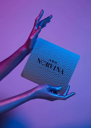 Two metallic painted hands holding a Norvina makeup palette against a purple and blue gradient background.