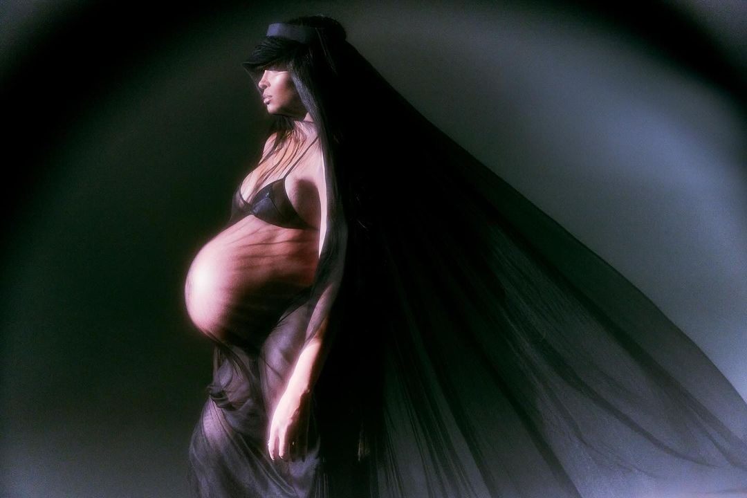 A pregnant woman in a black bra and sheer fabric poses in a photo studio with dramatic lighting and a dark background.
