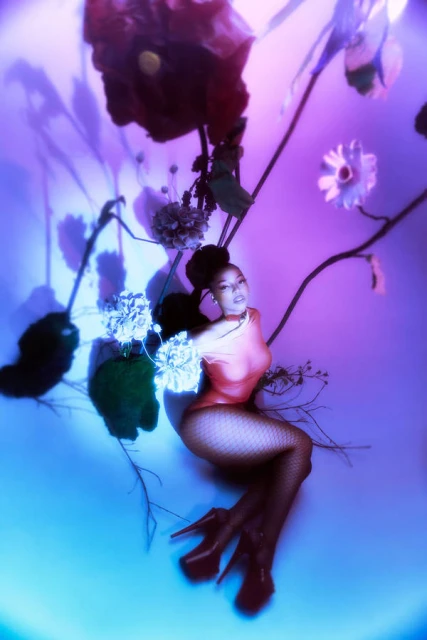 A young woman poses indoors with large dark red and white flowers. She is wearing a peach-colored bodysuit, high heels, and fishnet stockings.