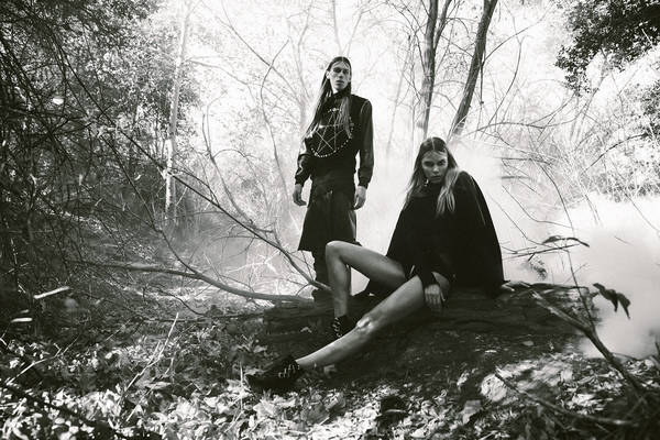 Monochrome image featuring an adult female seated on a log and an adult male standing in a wooded area filled with a light fog.