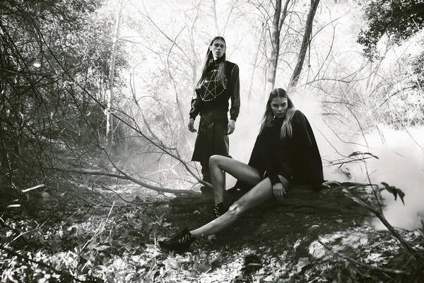 Monochrome image featuring an adult female seated on a log and an adult male standing in a wooded area filled with a light fog.