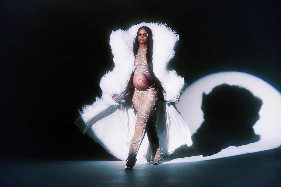 A pregnant woman in a sheer, embellished outfit and feathered coat poses in a spotlight against a dark background.
