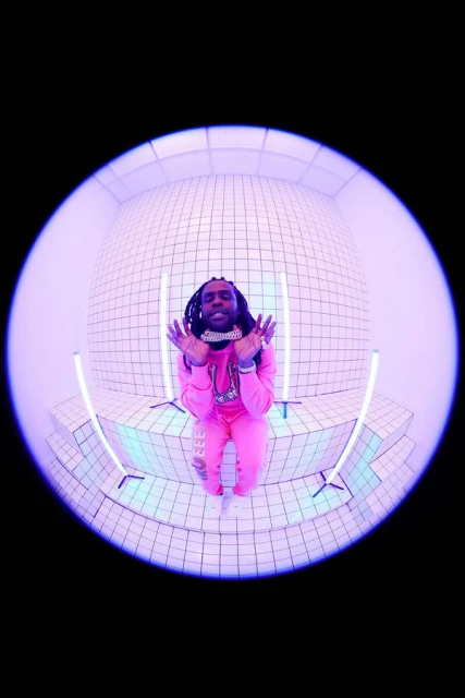 Young adult with dreadlocks poses in pink sweatsuit and jewelry against a tiled wall with neon lights, captured in a fisheye lens.