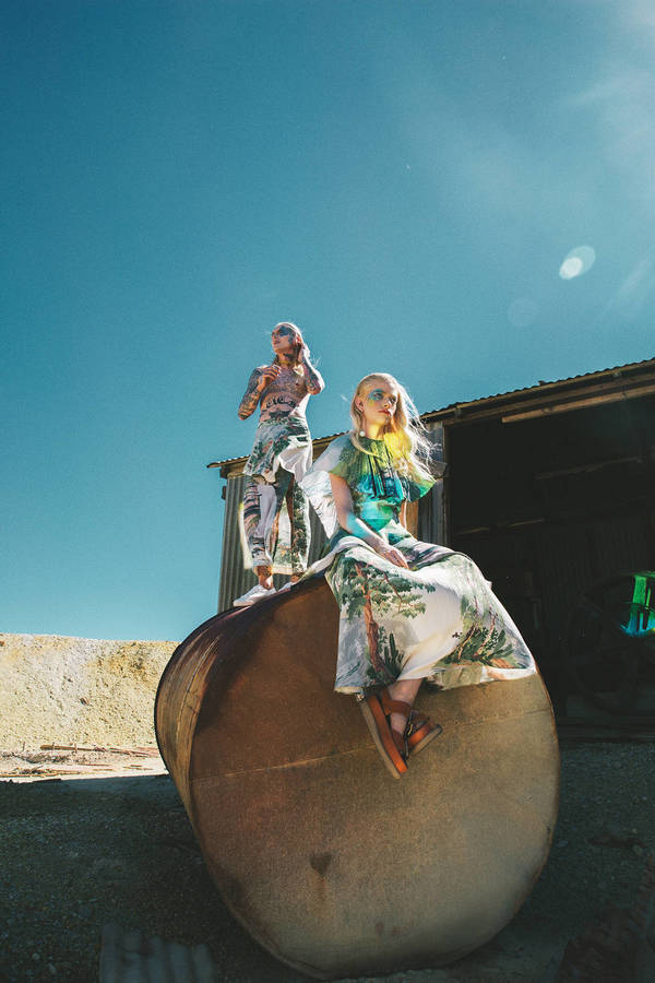 Two women in patterned clothing pose on a metal structure outdoors under a blue sky, creating a fashion editorial image.