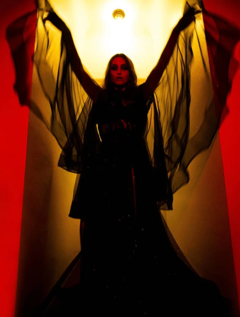 Woman in dark dress with arms raised dramatically, illuminated by upward lighting against a warm-toned wall, creating a stage-like effect.