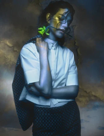 Portrait of a woman wearing a white collared shirt and dark patterned jacket with a green flower tucked behind her ear.