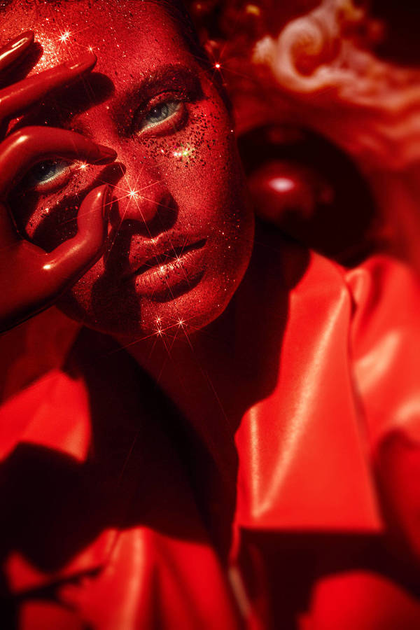 Close-up portrait of an adult female with glitter makeup, red face paint, and light eyes. She wears a red shirt and gestures with her hand.
