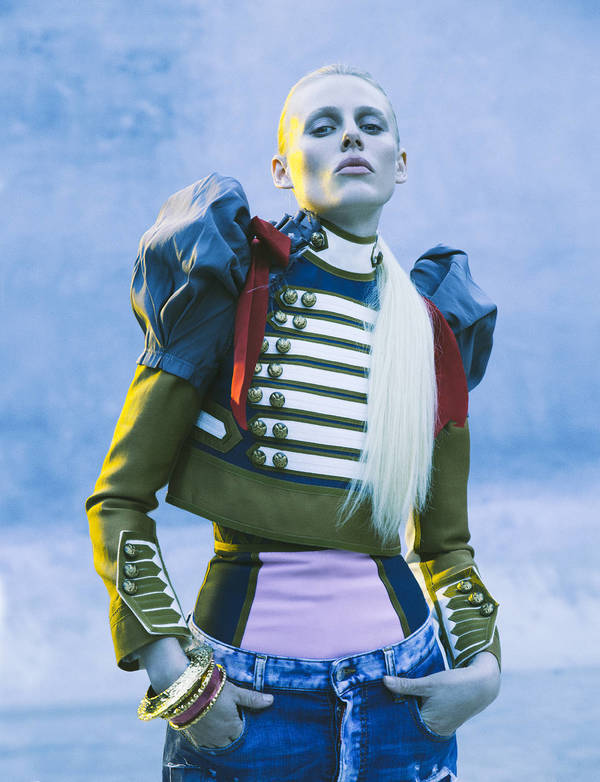 Stylish woman in cropped military-style jacket and jeans posing against a muted blue backdrop with gold bracelets.