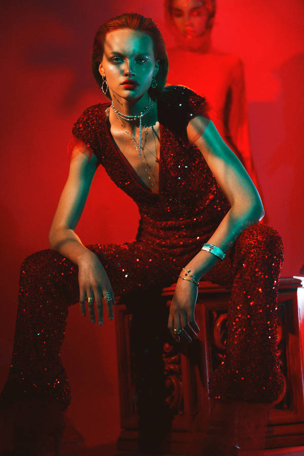 Glamorous young woman with long brown hair, wearing a red sequined jumpsuit, and silver jewelry, sitting on a pedestal in a red-lit studio.