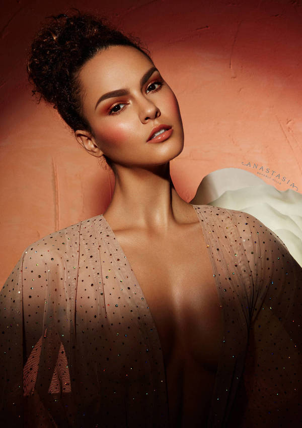 Portrait of a young adult woman with bronze eyeshadow, blush, and lipstick, wearing a sheer polka-dot top in front of a mottled peach background.