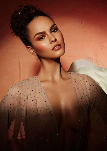 Portrait of a young adult woman with bronze eyeshadow, blush, and lipstick, wearing a sheer polka-dot top in front of a mottled peach background.