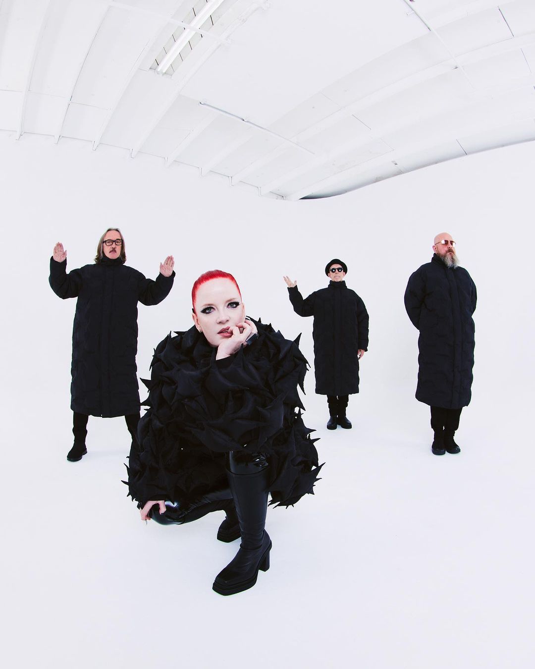 Four figures dressed in black stand in a white room, with a red-haired person kneeling in front in an avant-garde feathered coat.