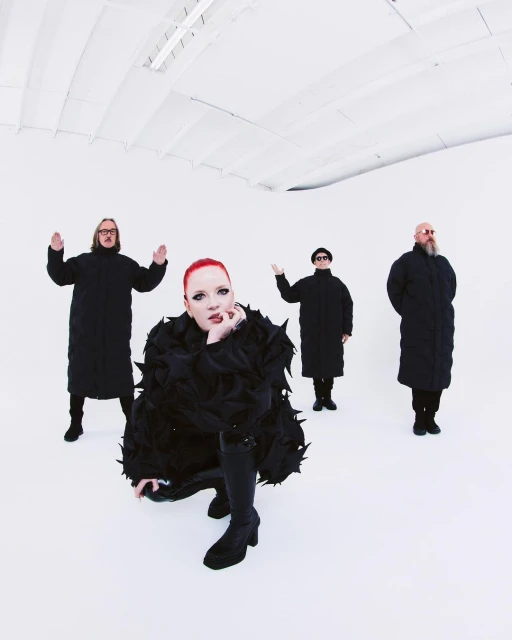 Four figures dressed in black stand in a white room, with a red-haired person kneeling in front in an avant-garde feathered coat.