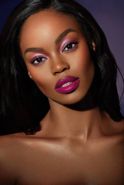 Close-up of a woman with pink eye shadow and glossy lipstick, highlighted cheekbones, and dark hair against a purple backdrop.