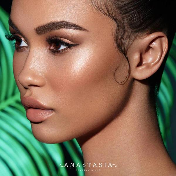 Close-up beauty shot of a woman with neutral makeup and glossy lips against a tropical green background, featuring Anastasia Beverly Hills logo.