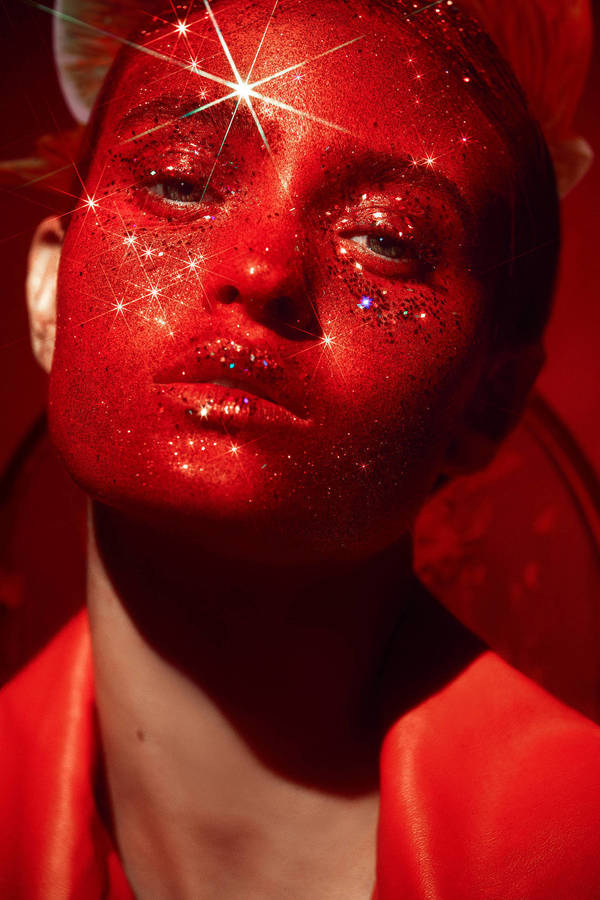 Portrait of a woman with red glitter makeup, a starburst lens flare, and a red leather jacket, set against a matching red background.