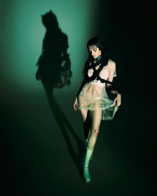 A woman in a sheer dress and chunky shoes stands in a studio with a shadow of horns behind her, set against a gradient backdrop.