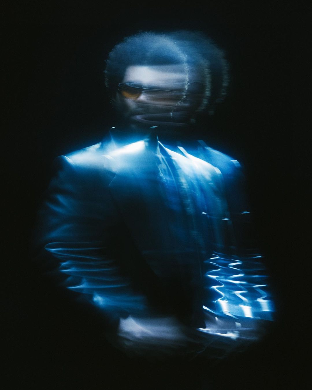 Portrait of male adult with sunglasses, wearing a suit, captured with intentional blur and a dark background, blue color cast.