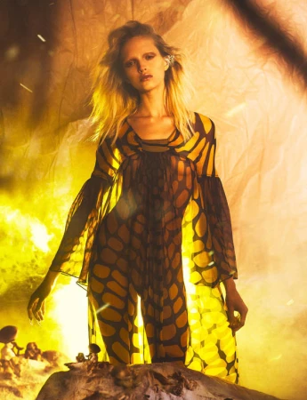 Fashion image of a fair-skinned adult female posing in a translucent jumpsuit with brown and yellow geometric patterns, set against a warm-toned backdrop.
