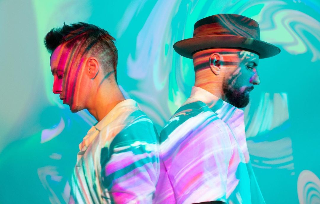 Two men stand back-to-back in a studio. One wears a hat, with patterned light projections on their faces and patterned shirts.