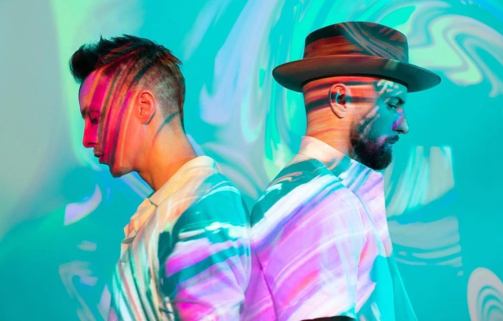 Two men stand back-to-back in a studio. One wears a hat, with patterned light projections on their faces and patterned shirts.
