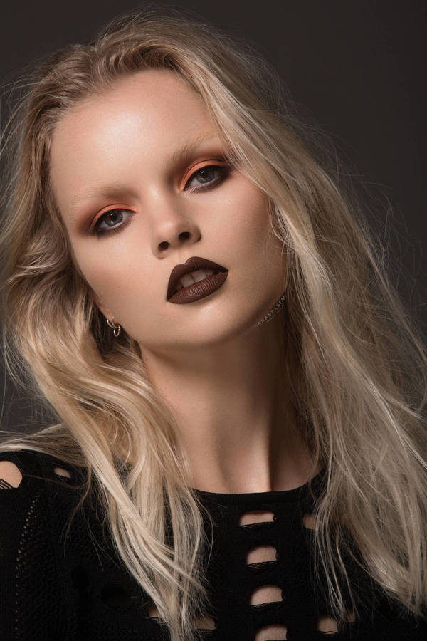 Close-up studio shot of a fair-skinned adult woman with blonde hair, dark lipstick, and orange eye makeup, against a dark gray background.