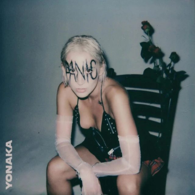 Indoor portrait of a young adult woman with 'PANIC' written across her face, wearing a studded black top and sheer gloves, seated in a chair.