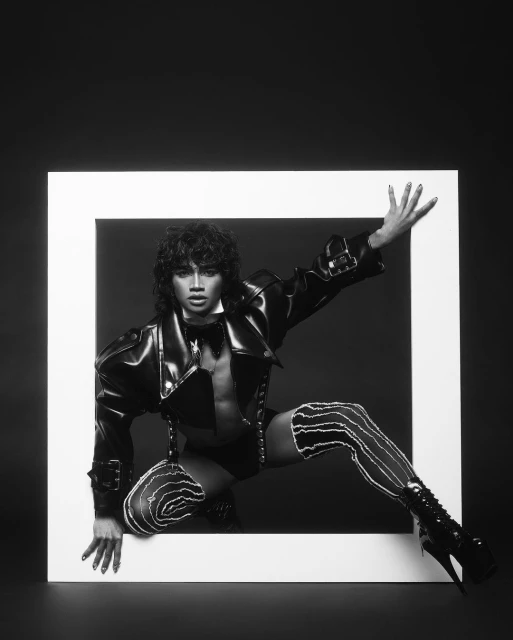Monochrome studio portrait of a person in a black patent leather jacket, shorts, bow tie, and striped stockings, framed in a square.
