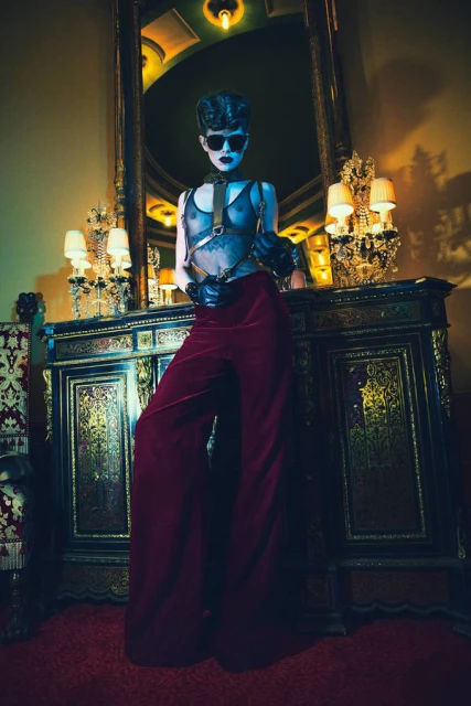 Stylish person stands in a luxurious room wearing wide-leg pants, a see-through top, and sunglasses, posing next to an ornate cabinet.