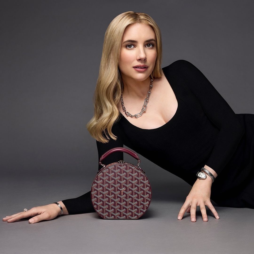 Emma Roberts poses with a patterned burgundy purse in a studio setting, wearing a black long-sleeve dress and silver jewelry.