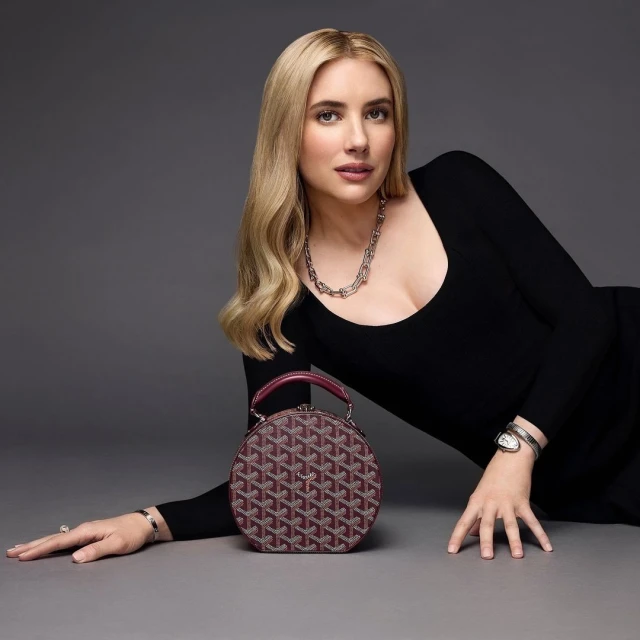 Emma Roberts poses with a patterned burgundy purse in a studio setting, wearing a black long-sleeve dress and silver jewelry.
