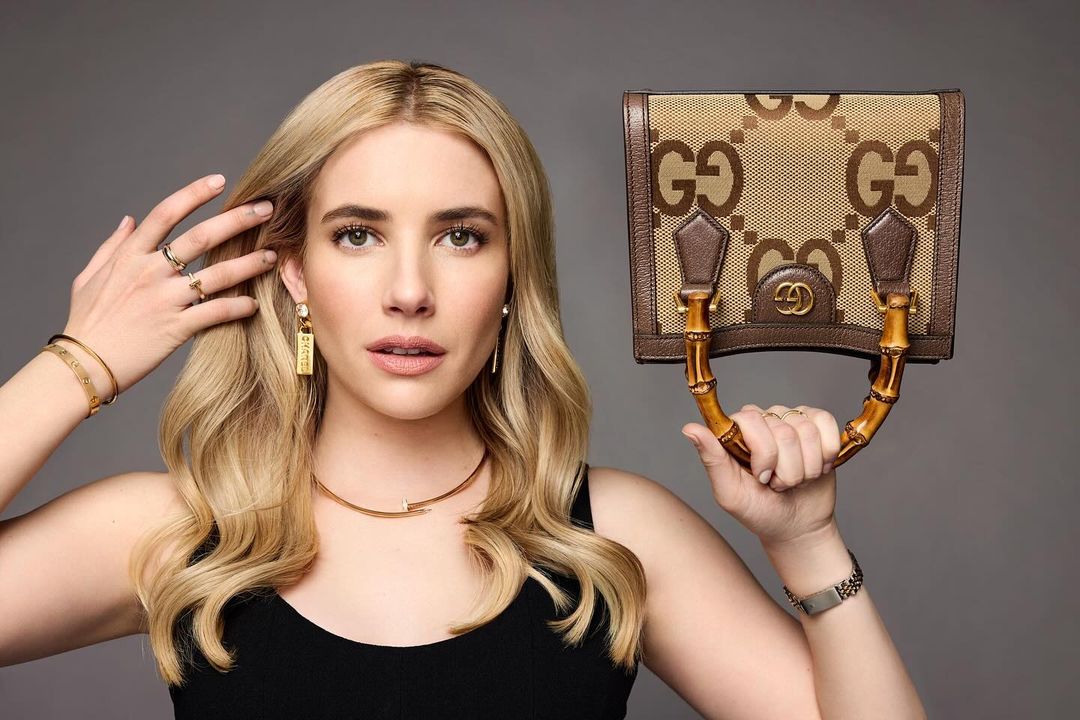 Emma Roberts poses with a Gucci bag in a studio shot, wearing gold jewelry against a gray backdrop. Fashion, luxury, and celebrity.