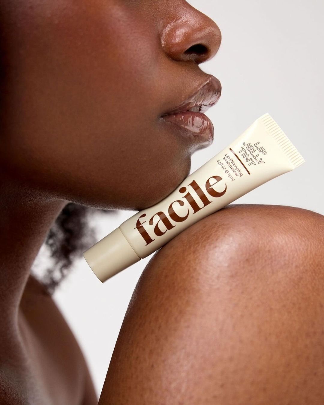 Close-up of a dark-skinned adult woman with glossy lips, posing with a tube of lip jelly tint from Facile against a white background.