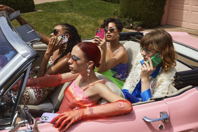 Four women sitting inside a vintage pink convertible car holding smartphones