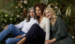 Three adult women of different ethnicities pose together in a studio setting surrounded by foliage, showcasing diverse beauty and friendship.