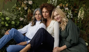 Three adult women of different ethnicities pose together in a studio setting surrounded by foliage, showcasing diverse beauty and friendship.