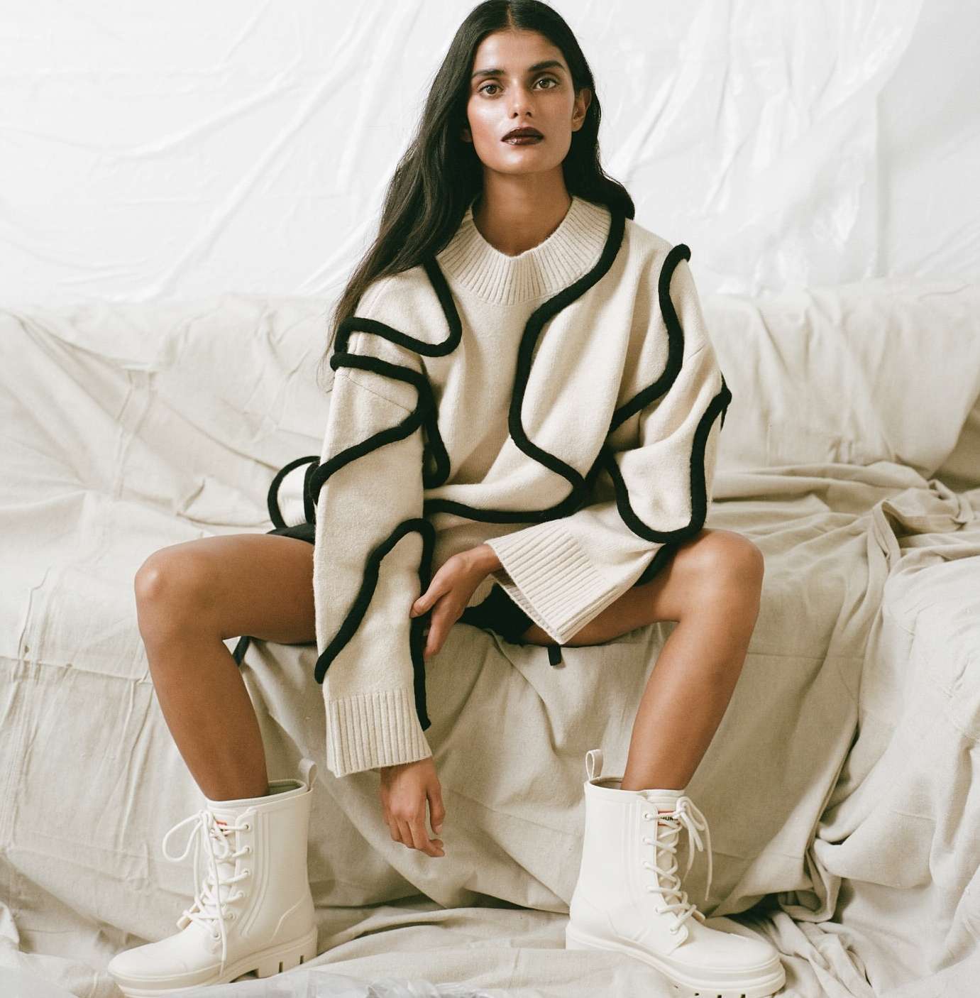 Woman sits on a bed wearing a beige sweater with black trim and matching boots against a neutral background, posing for a fashion shot.