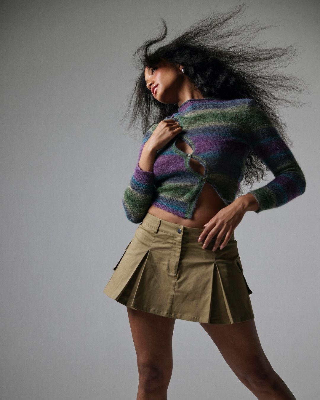 Woman with long dark hair blowing in wind wearing a striped cardigan and pleated mini skirt against a grey background.