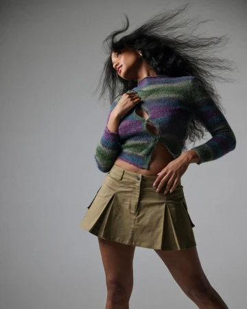 Woman with long dark hair blowing in wind wearing a striped cardigan and pleated mini skirt against a grey background.