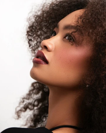 Close-up portrait of a woman with dark curly hair and deep burgundy lipstick looking upward against a white background.