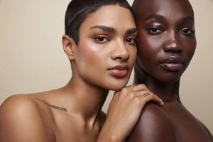 Close-up portrait of two female models with short hair posing together against a neutral cream background.