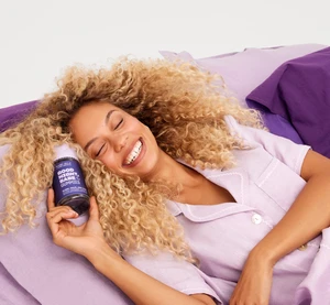 Smiling woman with curly blonde hair wearing light purple pyjamas holding a supplement bottle on a purple bed.