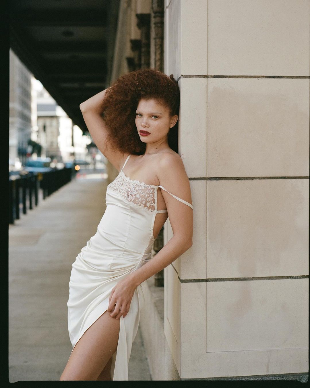 Young woman with afro poses in a dress on a city sidewalk leaning against a building. She has a slit in the dress showing her leg.