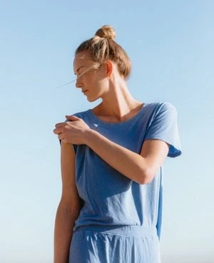 Woman in a blue dress poses against a light blue sky in an eye-level shot. Her hair is in a bun and she looks away from the camera.