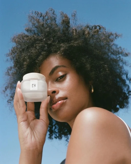 Woman poses with a cosmetic jar against a clear blue sky, showcasing beauty and skincare with a focus on natural light and product presentation.