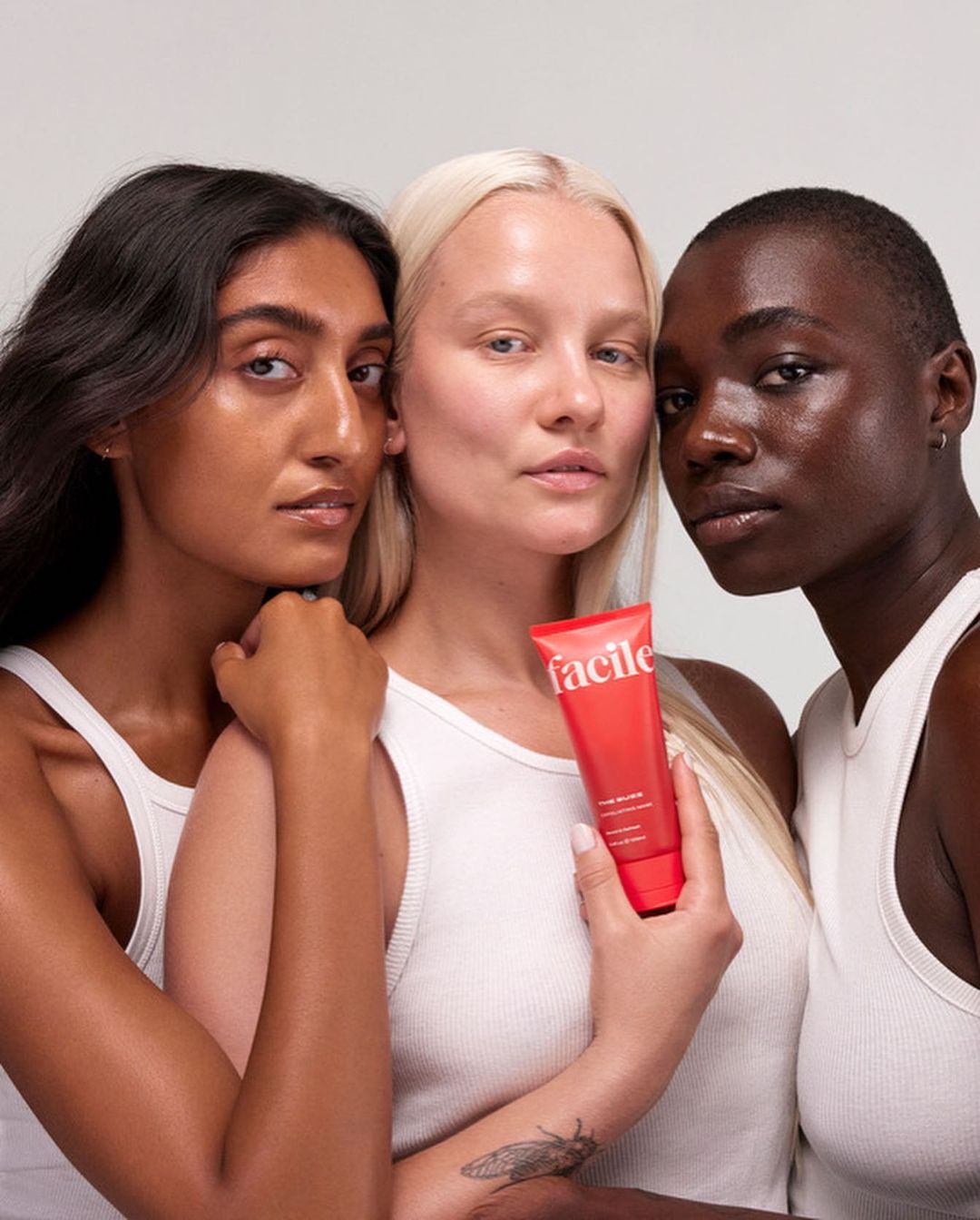 Three diverse women in white tops pose indoors holding a red Facile skincare tube against a neutral background in a beauty product shot.