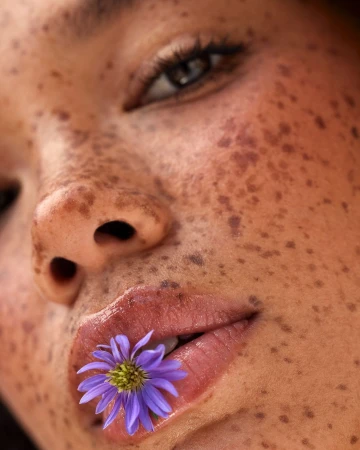 Extreme close-up portrait of a woman with heavy freckles holding a small purple flower between her lips.