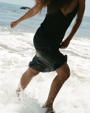 Woman in a black dress splashing through shallow ocean waves at the beach, photographed by Claire Leahy